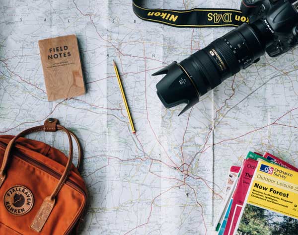 Flat lay of camera, backpack, notebook, and pencil on a map.