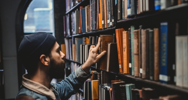 A Man wearing a jean jacket and beanie browses bookshelves in a library. 