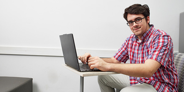 A student sitting at a small table desk using a laptop and smiling at the camera.