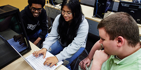 Three students sitting together at a desk in a computer lab, looking at a white laptop.