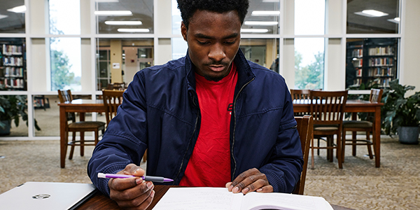 MGA student sitting at a table in the library looking down at a notebook with a pen in hand.