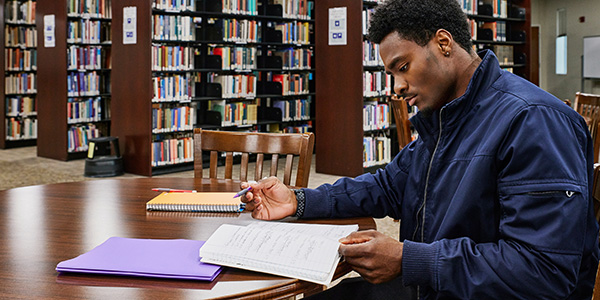 Student sitting at a table in a library looking down at a notebook with a pencil in his hand. 