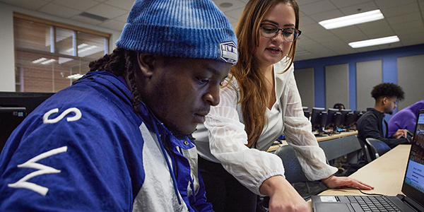 A staff member assists a student as they work together on a laptop in a computer lab classroom.