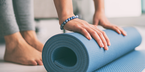 A close-up shot of a person's hands unrolling a blue yoga mat on a floor, with their bare feet visible in the blurred background.