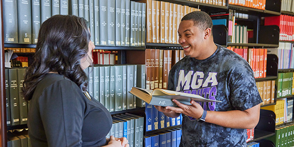 Image of a student smiling at a librarian beside shelves of book. The student is holding open a book in front of him.