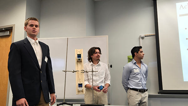 Three male students in professional attire present a physical project model next to a projection screen during a STEMposium session.