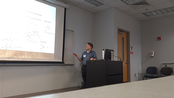 A male student stands at a podium and uses a clicker to present a slideshow featuring chemical diagrams and scientific data to an audience.