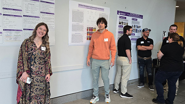 Two students stand in front of their research posters, including one titled "Artificial Sweeteners as Bacterial Energy Sources," while other attendees converse in the background.