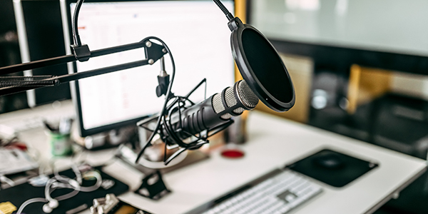 Image of a podcast setup, with a microphone and desktop computer sitting on a white desk.
