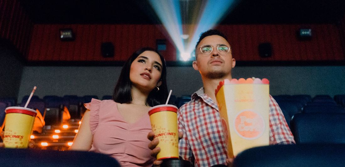 A man and woman sit in a dark theater holding popcorn and drinks watching a movie.