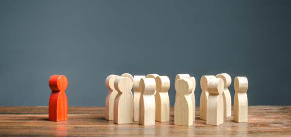 Wooden figurines stand on a flat surface. One red figure stands alone, separated from the natural colored figures. 