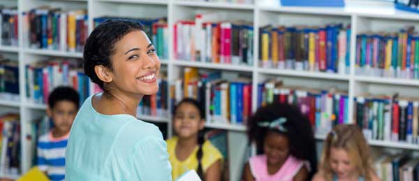 Teacher holding a book smiling in front of a classroom of students. 