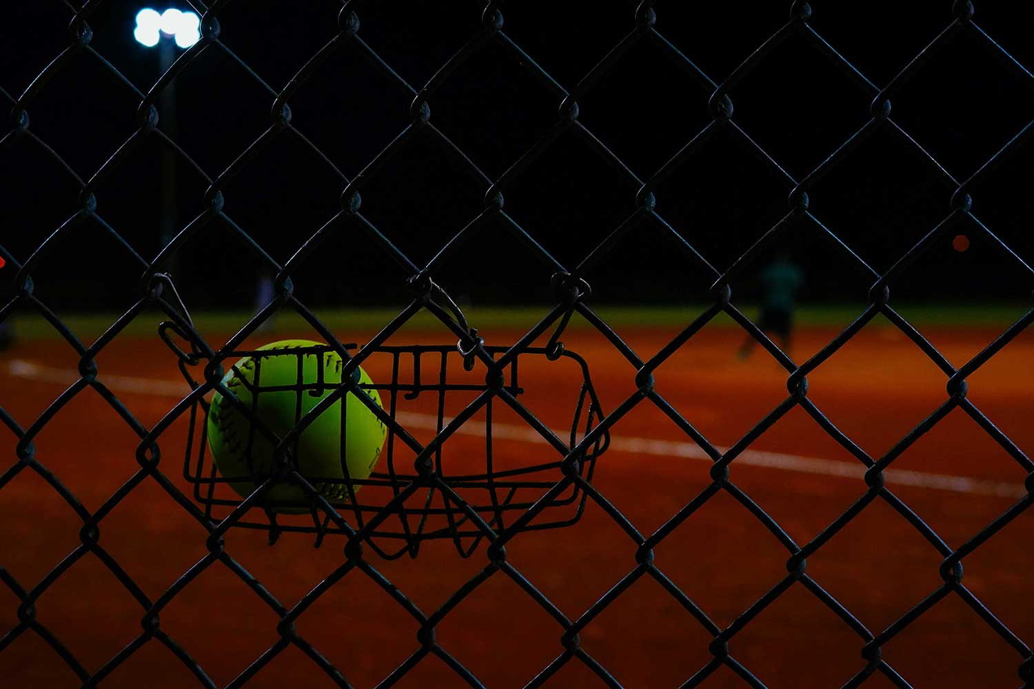 Softball sits in a basket hanging from a fence on a field.