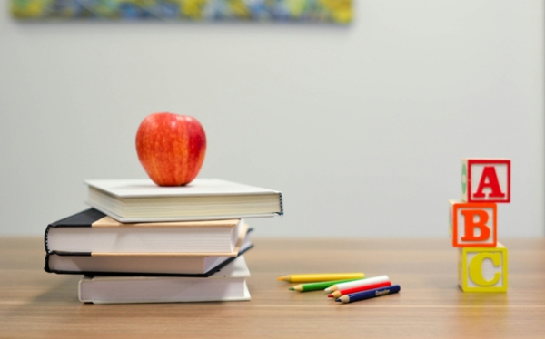 Apple, books, and stationary sit on a teacher's desk in a classroom.