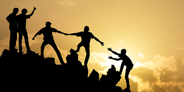 Silhouettes of a team helping each other climb a rocky peak against a golden sunset.