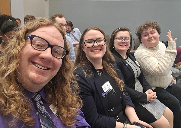A group selfie of the MGA Speech and Debate team members and their coach smiling in an auditorium.