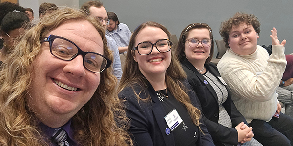A group selfie of the MGA Speech and Debate team members and their coach smiling in an auditorium.