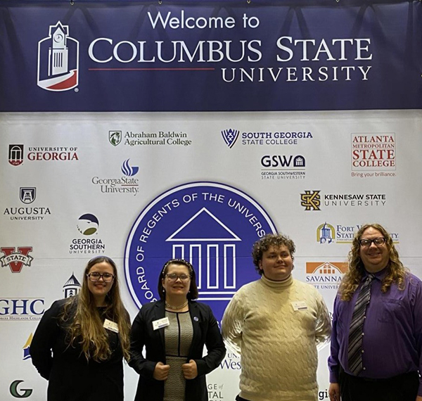 Members of the MGA Speech and Debate team and their coach standing in front of a welcome banner at Columbus State University.