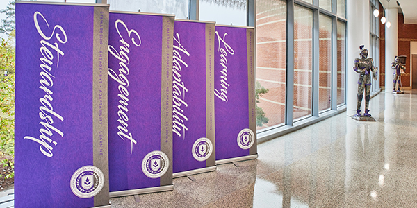 Four purple banners lined up in a university hallway displaying the MGA logo and the words Stewardship, Engagement, Adaptability, and Learning.