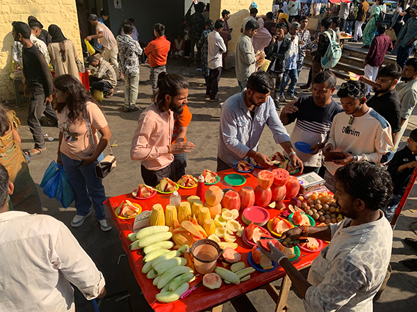 A street vendor in a crowded Indian market preparing fresh, sliced fruit at a bright red table for a group of people.