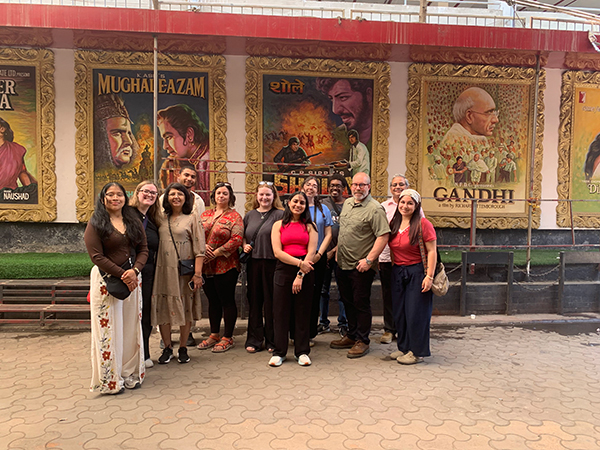 A group of MGA study abroad attendees and faculty posing outdoors in front of a wall featuring several oversized, colorful Bollywood movie posters.