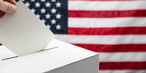 A vote being placed in a ballot box by someone off-screen with the American flag in the background.