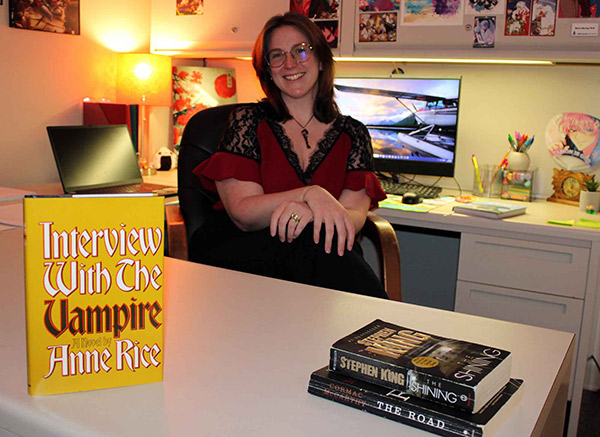 MGA professor, Dr. Storm Murray smiling and sitting at an office desk, with a hardback copy of "Interview With The Vampire" by Anne Rice standing upright in the foreground and a stack of books including Stephen King's "The Shining" and Cormac McCarthy's "The Road" beside it.
