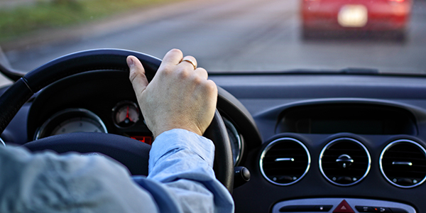 Image of individual with their hand on a steering wheel facing the road where another car is driving in front of them.