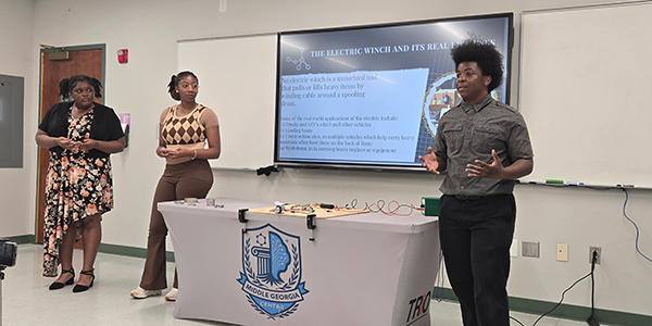 Three students presenting an electric winch project in front of a digital screen and a table with a Middle Georgia Center TRIO tablecloth.