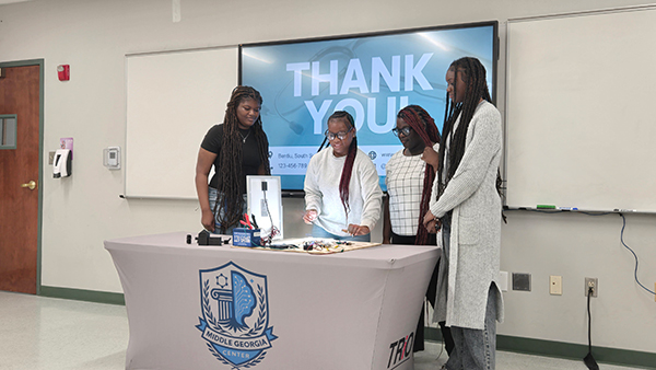 Four students examining a mini solar panel and circuitry setup on a presentation table with a Middle Georgia Center TRIO tablecloth.