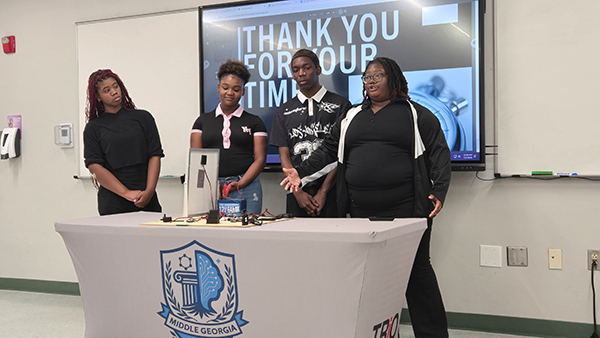 Four students concluding a presentation in front of a table with a solar panel project and a screen reading "Thank You For Your Time."