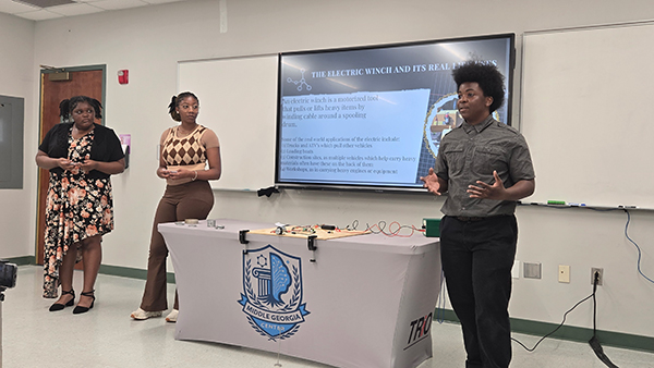 Three students presenting an electric winch project in front of a digital screen and a table with a Middle Georgia Center TRIO tablecloth.