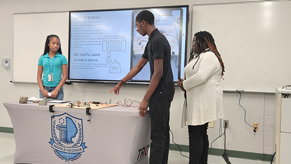 Three students demonstrating a STEM project in a classroom, pointing to equipment on a table with a Middle Georgia Center TRIO tablecloth.
